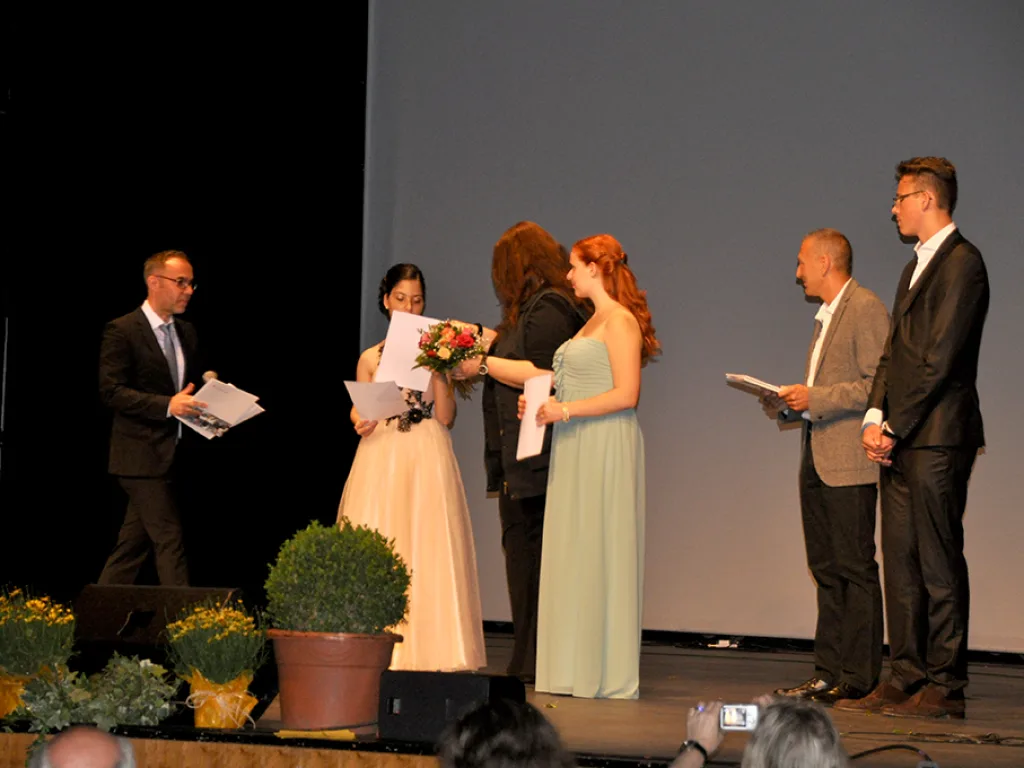 Zwei Absolventinnen nehmen zwischen drei Männern Blumen und Urkunden entgegen.